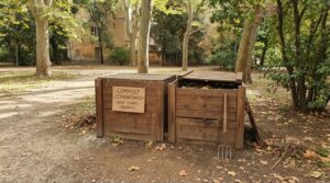 Isola di compostaggio comunitario in un parco urbano italiano con bidoni marroni e cartello scritto a mano